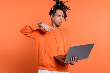 confused multiracial man with dreadlocks and tattoo pointing at laptop on coral background 