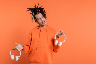 uncertain multiracial man with dreadlocks holding different wireless headphones and looking at camera on coral background 