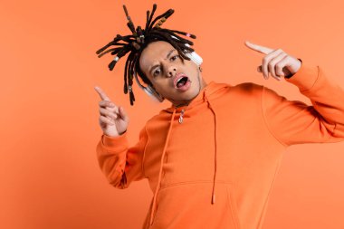 emotional multiracial man listening music in wireless headphones and pointing with fingers on coral background