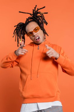 pierced multiracial man in hoodie wearing stylish sunglasses and pointing with fingers isolated on coral background