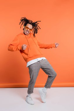 excited multiracial man in hoodie wearing stylish sunglasses and dancing on coral background