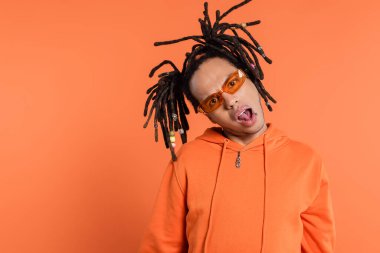 shocked multiracial man in stylish sunglasses looking at camera with opened mouth on coral background