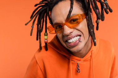 portrait of satisfied multiracial man in hoodie and stylish sunglasses isolated on coral 