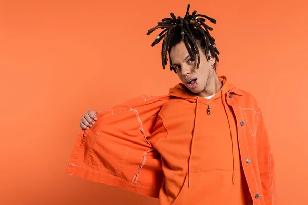 multiracial man with dreadlocks and opened mouth adjusting denim jacket on coral background
