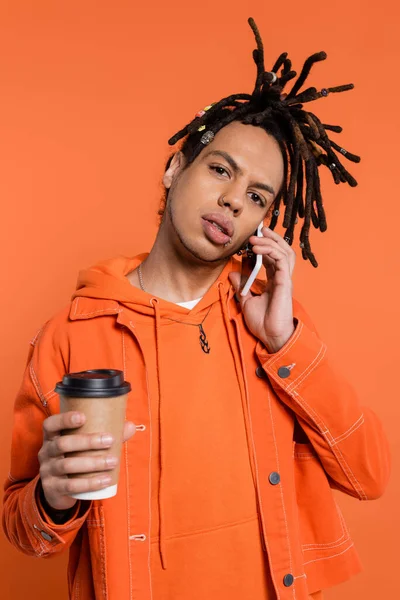 stock image pierced and multiracial man with dreadlocks holding paper cup and talking on smartphone isolated on coral 