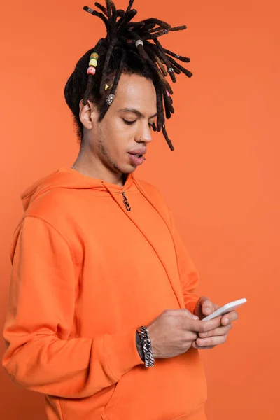 stock image multiracial man with dreadlocks standing in hoodie and texting on smartphone isolated on coral background 