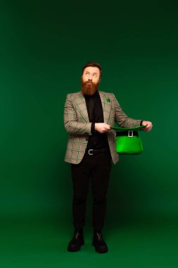Full length of bearded man in jacket holding hat during saint patrick day on green background 
