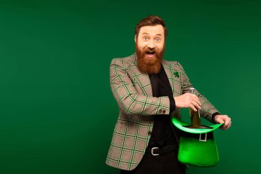 Excited bearded man holding hat and bottle of beer during saint patrick day isolated on green  clipart