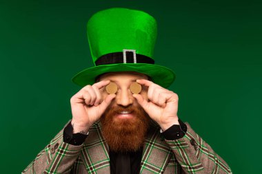 Smiling bearded man in hat holding coins near eyes isolated on green 