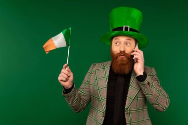Şapkalı sakallı adam akıllı telefondan konuşuyor ve yeşil üzerinde izole edilmiş İrlanda bayrağına bakıyor. 