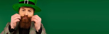 Portrait of man in hat touching moustache and looking at camera isolated on green, banner 