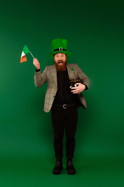 Full length of excited man in hat holding Irish flag and pot with coins on green background 