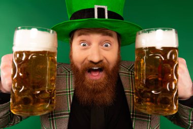 Portrait of excited man in hat holding beer glasses isolated on green 