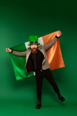 Full length of excited bearded man in hat holding Irish flag on green background 