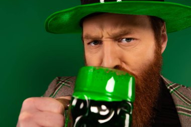 Portrait of serious man in hat drinking beer isolated on green  clipart