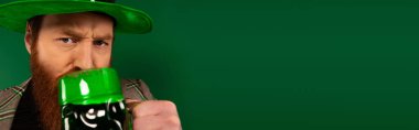 Serious man in hat drinking beer while celebrating saint patrick day isolated on green, banner   clipart