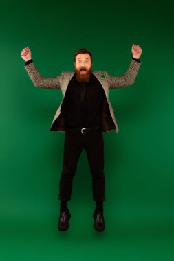 Excited bearded man in plaid jacket with clover jumping on green background 