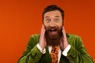 Trendy bearded model in jacket touching cheeks isolated on red 