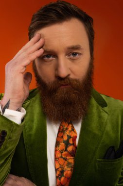 Portrait of stylish man in green jacket touching forehead isolated on red 