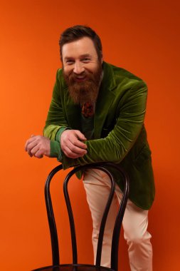 Smiling bearded model in jacket looking at camera near chair on red background 
