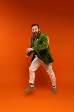 Cheerful bearded man in green jacket jumping on red background 