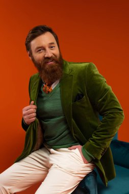 Smiling bearded man in stylish outfit sitting on armchair on red background 
