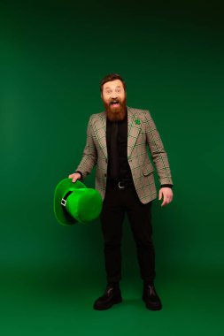 Excited bearded man holding hat and looking at camera during saint patrick celebration on green background 