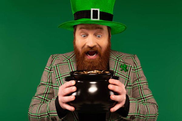 Amazed man in hat holding pot with golden coins isolated on green 