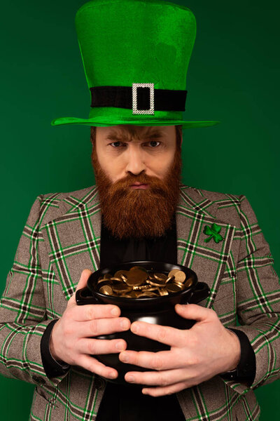 Angry man in hat with clover holding pot with golden coins isolated on green 