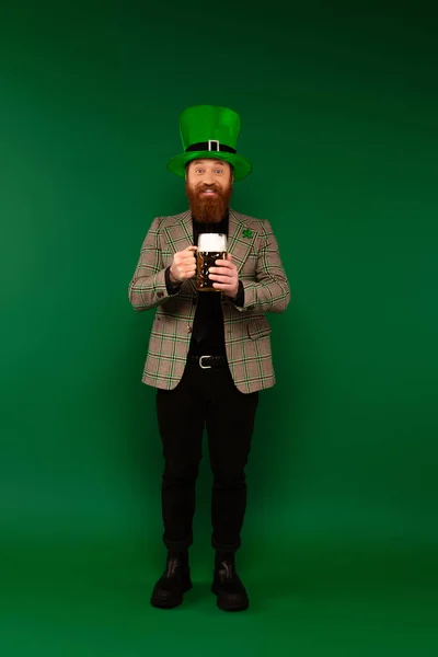 Stock image Full length of smiling bearded man in hat holding glass of beer on green background 