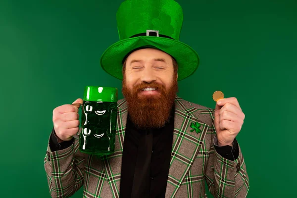 Pleased bearded man in hat holding beer and golden coin isolated on green 