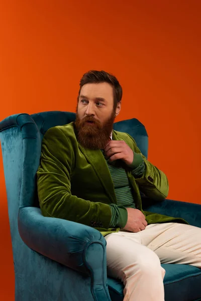 Fashionable bearded model sitting on velvet armchair on red background