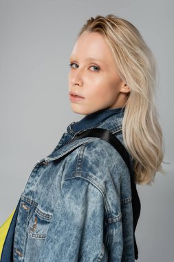 young blonde model in stylish denim jacket looking at camera while posing isolated on grey  clipart