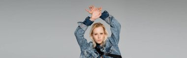 young blonde woman in stylish denim jacket looking at camera while posing with hands above head isolated on grey, banner  clipart