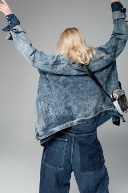 back view of young blonde model in stylish denim jacket with belt bag posing with raised hands isolated on grey  clipart