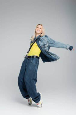 full length of happy young woman in blue denim outfit and stylish sneakers posing on grey 