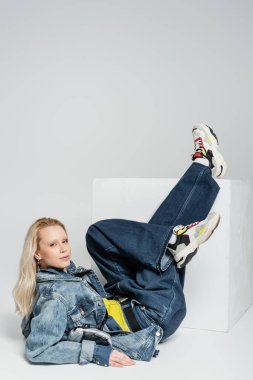 full length of blonde woman in blue denim outfit and trendy sneakers posing near white cube on grey  clipart