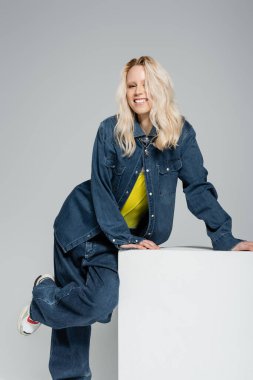 joyful young woman in blue denim outfit posing near white cube on grey 