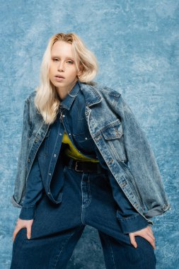 blonde woman in denim jacket posing while looking at camera near blue textured background  