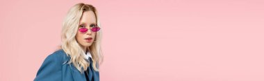 young blonde woman in blue blazer with tie and sunglasses posing with hands in pockets isolated on pink, banner  clipart