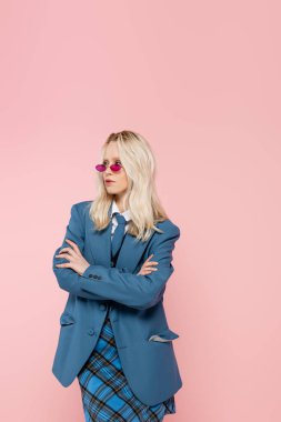 blonde woman in blue blazer and plaid skirt posing with crossed arms isolated on pink 