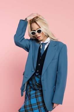 young woman in blue outfit and sunglasses adjusting blonde hair isolated on pink clipart