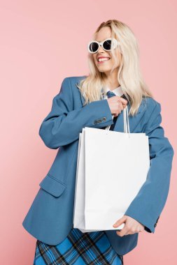 happy blonde woman in blue blazer and trendy sunglasses holding shopping bags isolated on pink clipart