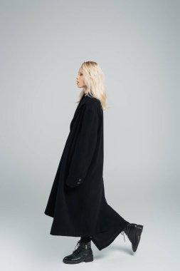 side view of blonde young woman in black coat walking and looking away on grey 