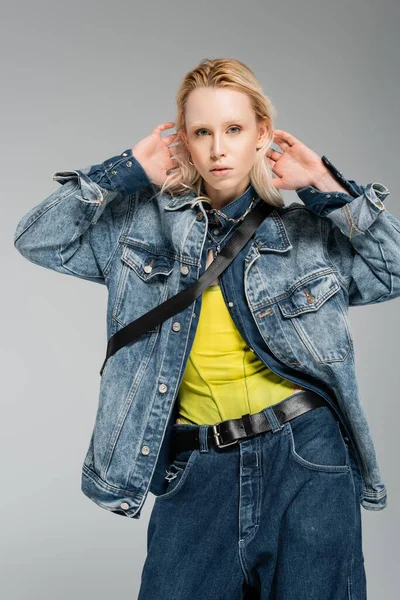 stock image young blonde woman in stylish denim outfit looking at camera while posing with hands behind head isolated on grey 