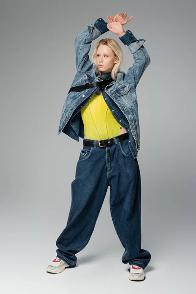 full length of blonde woman in stylish denim outfit posing with hands above head on grey