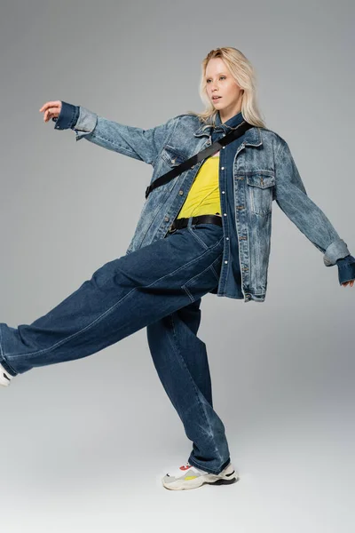 full length of blonde woman in stylish denim outfit posing while walking on grey