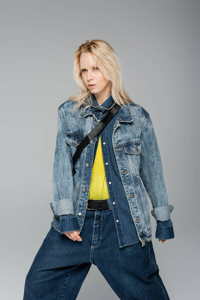 young blonde woman in stylish denim jacket posing while standing isolated on grey 