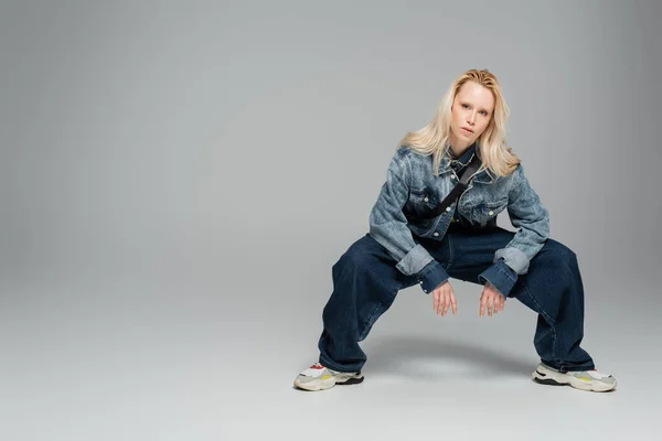 full length of young blonde woman in stylish denim clothes posing on grey
