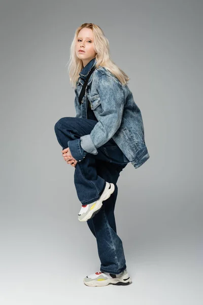 full length of blonde woman in trendy denim outfit and sneakers posing while standing on one leg on grey
